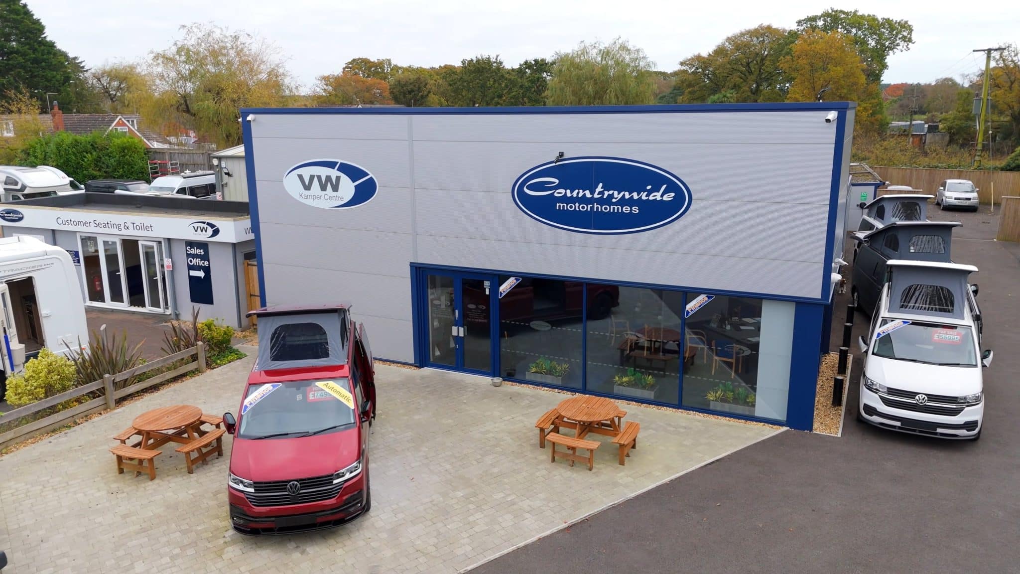 A red campervan with its roof popped up is parked outside a modern, grey and blue building with large windows. The building has "Countrywide motorhomes" and a "VW Van Centre" logo on its facade. Several other campervans and motorhomes are visible parked on the lot around the building, which is surrounded by trees under an overcast sky. Two wooden picnic tables are also in the foreground.