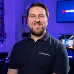 A person with short brown hair and a beard smiles while wearing a dark polo shirt with the southpoint films logo. They are in a media studio with blue lighting and a television screen displaying the company logo in the background.