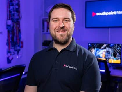 A person with short brown hair and a beard smiles while wearing a dark polo shirt with the southpoint films logo. They are in a media studio with blue lighting and a television screen displaying the company logo in the background.