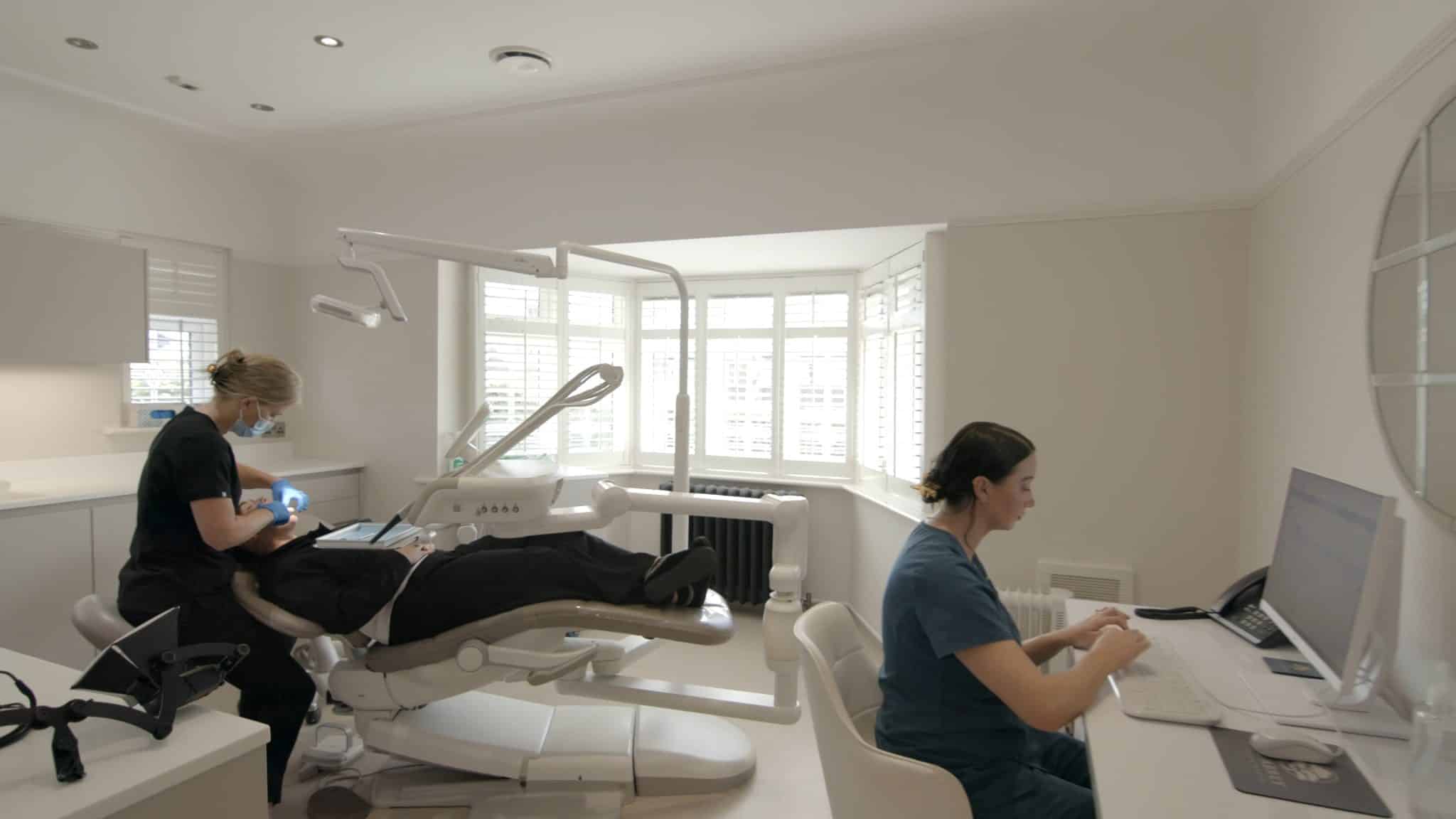 A modern, brightly lit dental examination room. On the left, a dental professional in blue scrubs and a mask is attending to a patient lying in a dental chair. On the right, another dental professional in blue scrubs is seated at a desk, looking at a computer monitor. Large windows with white shutters are visible in the background, letting in natural light.