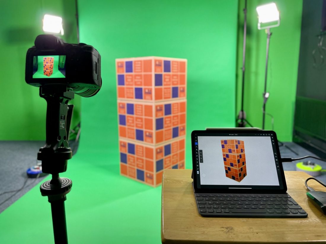 A photography studio. A stack of three cardboard boxes with Lokn'Store branding is placed in front of a large green backdrop. There are lights either side, a camera and an iPad running photo edited software present.