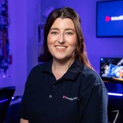 A person with long brown hair smiles while wearing a dark blue polo shirt with a logo, standing in a film studio with blue ambient lighting and a screen displaying "southpoint films".