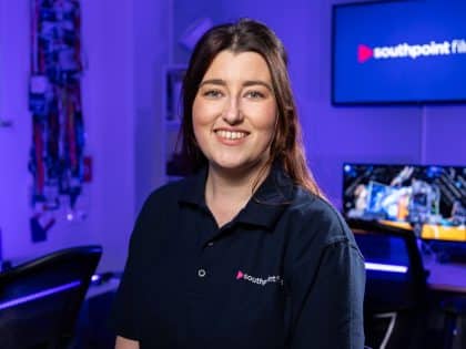 A person with long brown hair smiles while wearing a dark blue polo shirt with a logo, standing in a film studio with blue ambient lighting and a screen displaying "southpoint films".