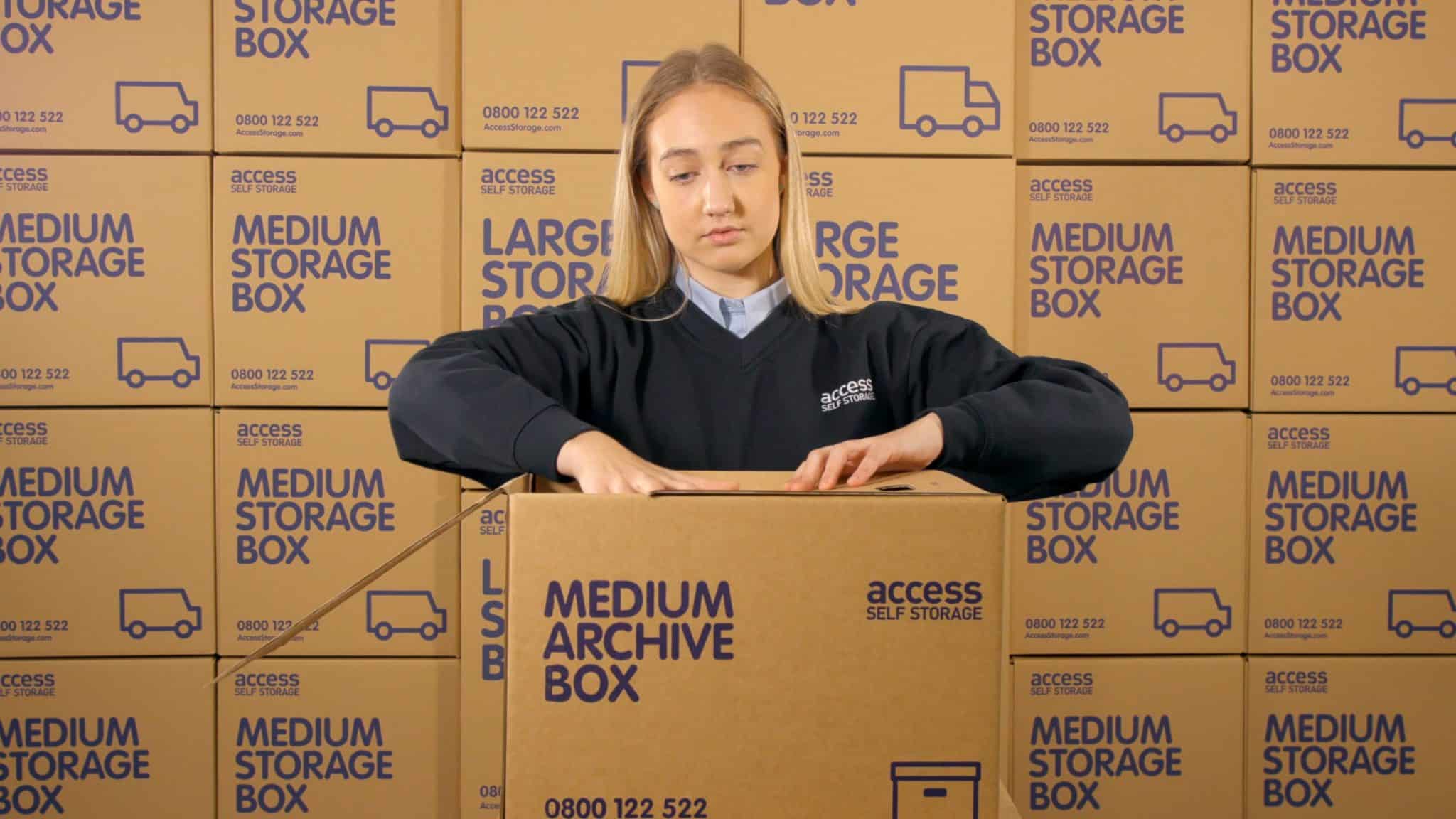 A woman in an "Access Self Storage" uniform is packing a box labeled "Medium Archive Box." She is surrounded by stacks of similar cardboard boxes, with the text "Medium Storage Box" and "Large Storage Box" printed on them. The boxes also display the company logo, phone number, and a graphic of a delivery truck. She is working in a storage or warehouse setting.