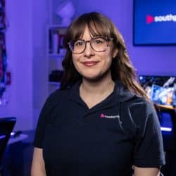 A person with long brown hair and glasses wearing a dark blue Southpoint polo shirt stands in a film studio with blue ambient lighting and a "southpoint films" logo displayed on a background screen.