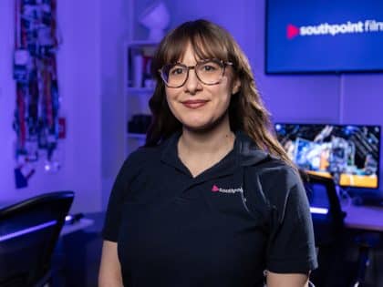 A person with long brown hair and glasses wearing a dark blue Southpoint polo shirt stands in a film studio with blue ambient lighting and a "southpoint films" logo displayed on a background screen.