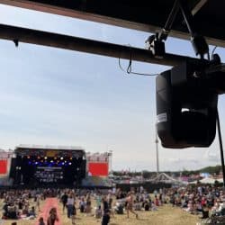 A close-up shot of a black PTZ camera mounted upside down on an overhead metal beam, with a large outdoor stage and a crowd of people in the background under a bright sky.