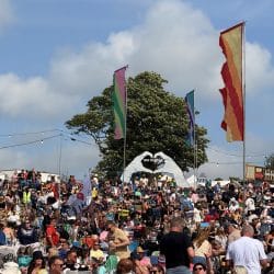 A wide shot of a large outdoor music festival or event with a densely packed crowd seated on a grassy hill. In the background, there's a large white tent heart-shaped structure, a prominent tree, and two tall flags in rainbow and green/blue colours. String lights are visible across the top of the scene.