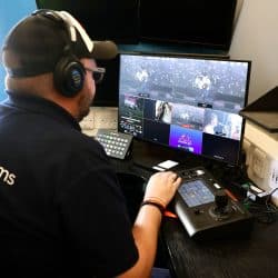 A person wearing headphones and a "southpoint films" polo shirt is seated at a desk, looking at a multi-screen computer monitor displaying multiple video feeds. Their hands are on a control panel with a joystick, indicating live video mixing or production.