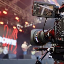 A close-up side view of a professional video camera on a tripod, with its monitor facing the viewer, positioned in front of a brightly lit outdoor stage. Red lights illuminate the stage, and blurry figures of musicians, one playing a guitar, are visible in the background. A large screen on the stage displays some text, part of which reads "UM BOWIE".