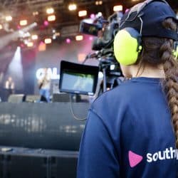 A person with braided hair, wearing yellow headphones and a "southpoint films" shirt, stands with their back to the viewer, looking towards a brightly lit stage with performers in the distance. A professional video camera on a tripod is in the foreground.