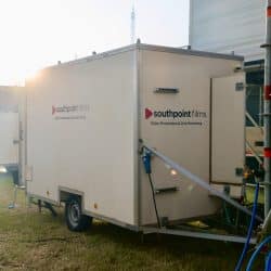 A white production trailer, bearing the "southpoint films" logo and "Video Production & Live Streaming" text, is parked on a grassy field. Sunlight creates a lens flare on the side of the front trailer.