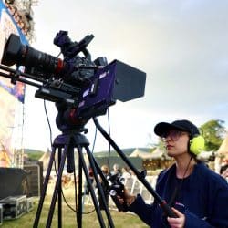 A person wearing a cap and yellow headphones stands next to a professional video camera on a tripod, looking intently towards a distant stage or area. They are wearing a blue "southpoint films" shirt.