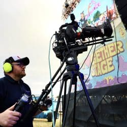 A person wearing a baseball cap and headphones stands operating a large professional video camera on a tripod, with a brightly lit stage featuring a colorful screen in the background. The person is wearing a "southpoint films" polo shirt.