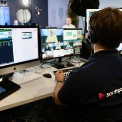 A technician wearing a "southpoint films" shirt and a headset sits at a desk managing multiple monitors during a live broadcast or recording. The screens show various video feeds, including live shots of speakers and presentation slides. Studio lights and cameras are visible in the background, with a person in a white shirt on set. The technician operates a control panel and keyboard, ensuring smooth transitions between video feeds.