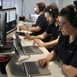 A team of three technicians wearing headsets work at a row of desks with laptops and monitors. The woman in the foreground types on a laptop, while the others focus on their screens or take notes. Multiple cables and equipment are visible around the workspace, suggesting they are managing a live broadcast or production. In the background, a man is seated, possibly being recorded or filmed.