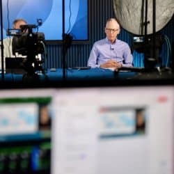 A man in a light blue shirt sits at a table, speaking on camera during a video production. Studio lights, cameras, and other equipment surround him, while another person partially visible sits beside him. The scene is framed by a blurred foreground showing a computer monitor with a live video feed. The man appears focused, possibly addressing a virtual audience or participating in a live broadcast.