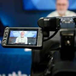 A video camera captures a man wearing glasses and a light green shirt as he speaks during a recording. The camera's display screen shows a clear image of the man, framed against a blue background, while the actual subject appears blurred in the background. The setting includes professional lighting and production equipment, with the man likely delivering a presentation or participating in a broadcast. The blue tablecloth partially visible reads "southern" as part of a larger logo.