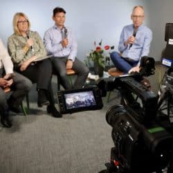 Four people sit in chairs holding microphones during a panel discussion, being recorded by a professional video camera in the foreground. The camera’s screen shows the group as they appear on film. The participants, two men and two women, are dressed in business casual attire, and a potted plant is positioned next to them. Cables and equipment are visible in the background, indicating a studio or broadcast setting. They appear to be engaged in conversation or answering questions.