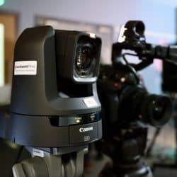 A close-up of a Canon PTZ camera mounted on a tripod, with a label that reads "southpoint films" and contact details on the side. In the background, another professional video camera is set up, along with lighting equipment, suggesting a video production or broadcast environment. The room appears to be a studio or a control room, with blurred monitors and equipment in the distance.