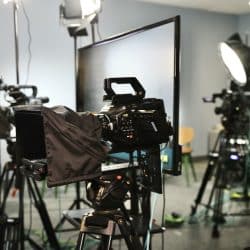 A professional video production setup with multiple cameras mounted on tripods, some covered with protective hoods, positioned around a large monitor. Bright studio lights and reflectors are arranged to illuminate the scene. Several cables run across the floor, connecting the equipment. The background includes more lights and production gear, indicating a controlled studio environment, likely for filming or live broadcasting.