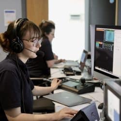 A woman wearing glasses and a headset sits at a desk, focused on a computer screen displaying a live video feed. She is operating a laptop and audio equipment, likely part of a production team. In the background, another woman is seated at a similar workstation, also working on a computer. The environment appears to be a control room for managing a live event or broadcast, with multiple screens and technical equipment visible.