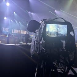 A backstage view of a concert with a focus on a camera capturing the performance. The camera screen shows a live feed of the stage, where "The Libertines" band name is displayed in large letters. Stage lights beam across the venue, highlighting amplifiers and a drum set in the background. The camera is covered with a protective plastic cover, suggesting it's being used in an outdoor or large-scale event.