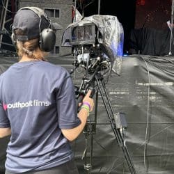 A camera operator stands in front of a stage, filming an event. The operator wears a headset and a navy "Southpoint Films" t-shirt, controlling a professional camera mounted on a tripod. The camera is covered with a protective plastic cover. On stage, a person dressed in black is partially visible near a microphone stand, with a backdrop featuring vibrant red and orange patterns. Various cables run along the floor, connecting equipment.