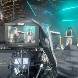 A close-up of a camera capturing a live performance on stage. The camera's screen shows two musicians, performing under teal stage lights. The camera is covered in a plastic protective sleeve, labeled "Southpoint Films." In the background, stage scaffolding, instruments, and additional lighting equipment are visible. Cables run along the floor, and the atmosphere is filled with a smoky concert ambiance.
