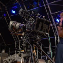 A person wearing large headphones operates a professional cinema camera on a tripod at a live event. The camera's monitor displays a live feed of a performer on stage under blue stage lighting.
