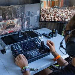 A person with headphones and several event wristbands operates a video switcher and joystick controller at a desk. Two monitors in front of them show multiple camera feeds of a large outdoor concert crowd and stage.