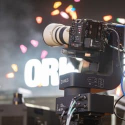 A professional cinema camera with a large white lens is mounted on a robotic ZANUS motion control head. The background is blurred with colorful stage lights and a large illuminated sign.
