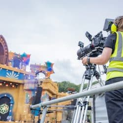 A person in a high-visibility yellow vest and ear protection operates a large camera on a tall tripod, overlooking an elaborate outdoor festival stage with circular floral motifs.