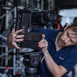 A person in a dark polo shirt carefully adjusts a cinema camera mounted on a ZANUS robotic head. The setup is located within a metal scaffolding structure.