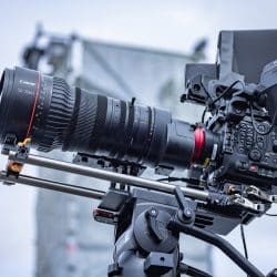 A close-up side profile of a professional cinema camera equipped with a massive 50-1000mm Canon lens and multiple monitors, mounted on a heavy-duty tripod.