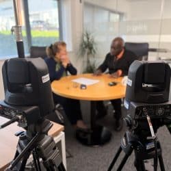 Two people are seated at a round table, facing each other, with camera and lighting equipment set up around them.
