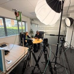 Two people are seated at a round table, facing each other, with camera and lighting equipment set up around them.