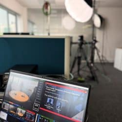 A computer running vision mixing software is visible in the foreground, with cameras and lights blurry in the background.