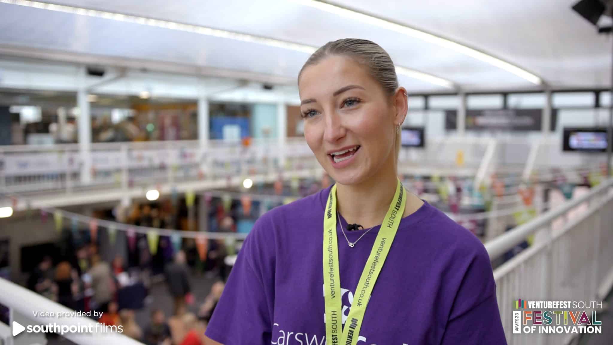 A woman wearing a purple shirt and yellow lanyard is speaking at an event. The lanyard has "venturefestsouth.co.uk" written on it. In the background, a busy venue with colorful decorations and people walking around is visible. The setting appears to be a tech or innovation festival. The bottom right corner has the "Venturefest South Festival of Innovation" logo, while the bottom left says, "Video provided by Southpoint Films."