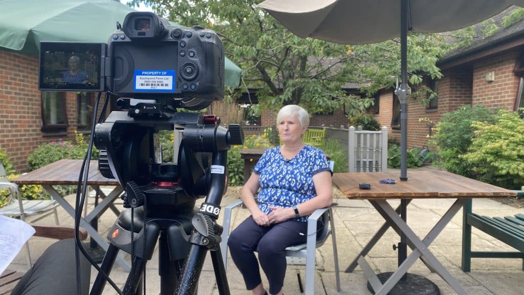 A professional camera mounted on a tripod is set up for an interview. In the background, a woman with short blonde hair wearing a blue patterned blouse is seated, looking slightly to the side. The setting is an outdoor patio with a wooden table, chairs, and a parasol. A garden and a brick building can be seen in the background.
