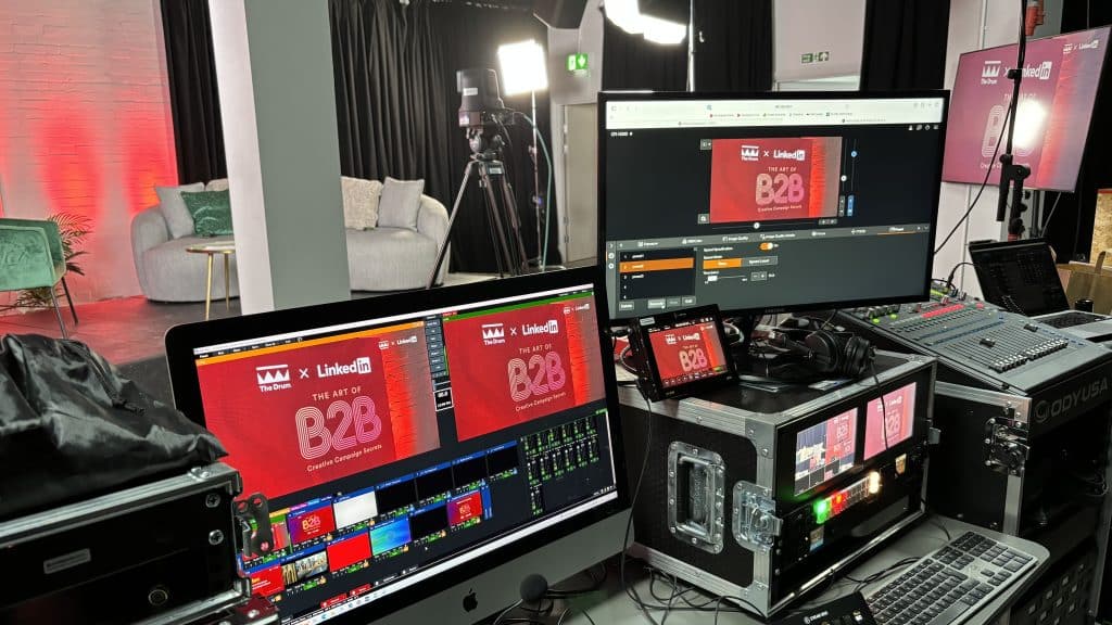 A behind-the-scenes view of a live broadcast setup featuring multiple monitors with graphics for "THE ART OF B2B" sponsored by LinkedIn and The Drum. A studio environment with professional lighting and a video camera on a tripod is visible in the background, highlighting a set with a grey couch and red-lit wall. The foreground shows production equipment, including a sound mixer and various computer interfaces, indicating a high level of technical control for live streaming or recording.