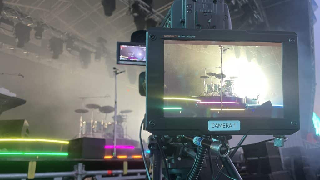 A camera's digital monitor labeled "CAMERA 1" shows a live feed of a drum set on a stage, bathed in dramatic lighting and stage fog. The viewpoint is from behind the camera, capturing part of the equipment and the broader stage setting, which includes colorful lighting effects. The scene suggests a music concert setting, with the focus on the drummer's area during a performance or sound check.