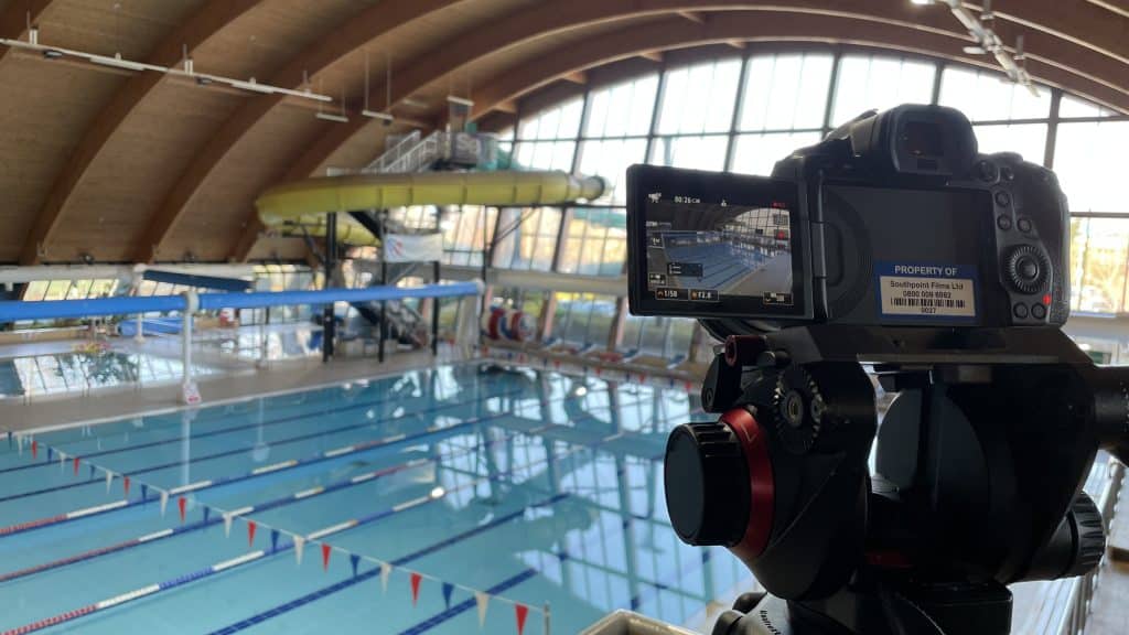 A camera on a tripod is aimed at an indoor swimming pool, with the camera's screen showing a view of the pool lanes. The backdrop features a large, wood-beamed ceiling and a waterslide, conveying a leisure or sports centre atmosphere. The pool is marked with lane dividers and the calm water reflects the structure above.