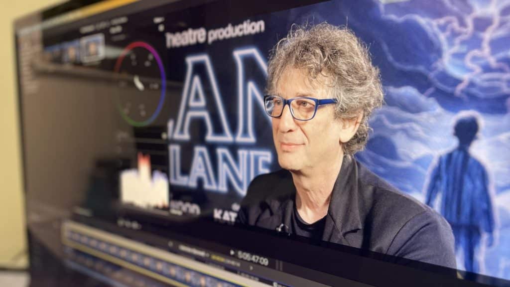 On a computer screen, video editing software is open with a timeline visible. An interview subject, a man with curly hair wearing glasses and a black shirt, is in focus on the preview monitor. The background of the video features a stylised, blue-hued image of a figure standing in a landscape, for a theatre production titled "Ocean at the End of the Lane". The setting suggests a post-production editing workflow for media content.