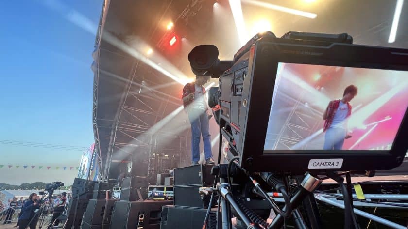 A live concert scene viewed from the side of the stage, showing a performer in mid-motion on stage, with the audience in the background. Stage lights cast bright spots across the scene. In the foreground, a professional video camera on a tripod is focused on the performer, with the camera's monitor displaying a live feed of the performance. The environment suggests an outdoor festival during dusk or early evening.