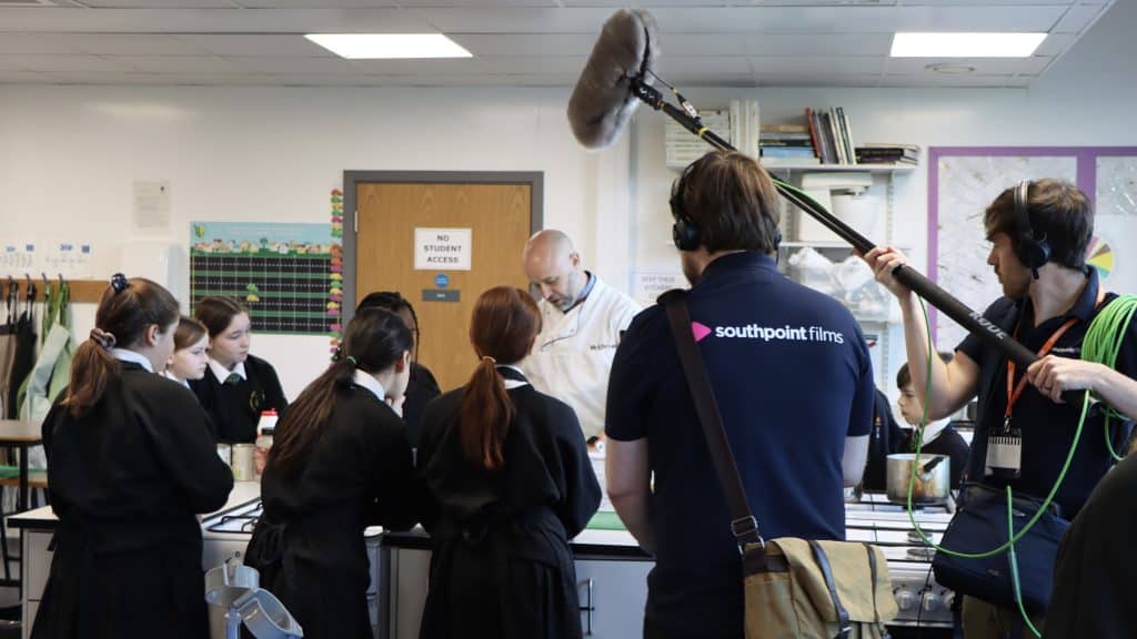A classroom scene where a group of students in uniforms are gathered around a chef demonstrating a cooking technique. In the foreground, a film crew from Southpoint Films is recording the session. One crew member is holding a microphone boom over the group, and another is wearing headphones and operating the camera (which we cant see).