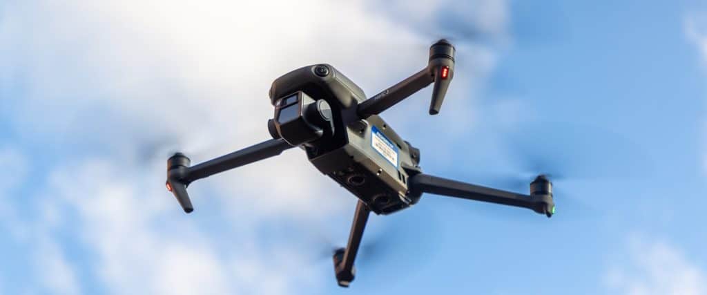 A black quadcopter drone with a camera, labeled "MAVIC", is captured in mid-flight against a soft-focus blue sky with light clouds. The drone's rotors are in motion, indicated by a slight blur, and red and green navigation lights are visible on the tips of its arms.