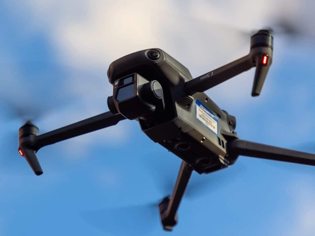 Close-up of a dark grey DJI Mavic 3 drone in flight, captured from below against a soft blue sky with blurred white clouds. The drone's propellers are spinning, creating motion blur, and red navigation lights are visible on its arms. The drone's camera and sensors are facing forward, ready for aerial photography or videography.