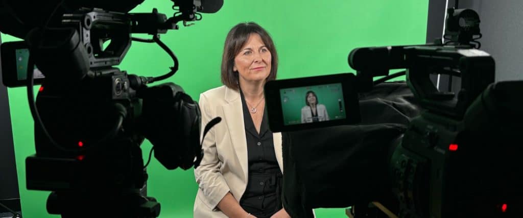 A woman in a beige blazer and black shirt is seated and smiling slightly towards the camera during a video production, with a green screen background. Two professional video cameras are focused on her, one displaying her image on its screen, illustrating a behind-the-scenes view of a broadcast or filming setup.