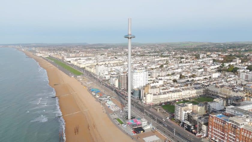 Aerial view of a coastal cityscape with a prominent tower in the center, overlooking a long sandy beach. The urban area is densely packed with buildings, and a clear demarcation between the beachfront and the city can be seen. The ocean is calm, with gentle waves lapping the shore, and the city extends into the horizon where gentle hills can be observed in the distance.